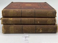 Side view of three stacked hardcover books showing worn spines with gold title text and embossed decoration.