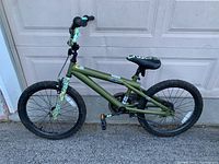 Front angle view of green kids bike with camouflage bike frame parts and seat against garage door background, showing side profile and tires.