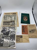 Photo showing a collection of vintage Eaton paper ephemera including advertisements for coats, shoes, men’s pants, and a 'Recollections' poetry booklet.