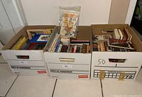 Three large banker-type boxes filled with assorted books, showing a mix of hardcovers and paperbacks packed tightly within.