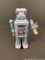 Front view of small blue wind-up tin robot showing detailed painted front panel, red eyes, and sided arms; minor scratches visible on surface.