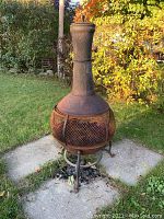 Outdoor photo of a large metal chiminea showing significant rust damage and wear, placed on a concrete pad in a grassy yard with fall foliage in the background.