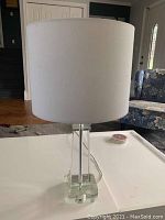 Side view of the clear acrylic lamp with white drum shade on a white table.