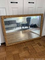 Full view of the large rectangular mirror with gold ornate frame leaning against white doors on wooden floor, reflecting living room furniture.