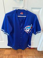 Front view of the blue Toronto Blue Jays jersey hanging showing full front with buttons and logo patch.