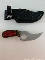 Curved blade knife with finger hole and wooden handle next to black sheath with belt clip