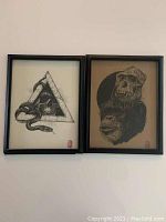 Two framed black and white art prints side by side. One features a snake and skull in a triangle; the other displays a gorilla skull with a dark circular background. Both prints have red artist marks in the corner. One of the frames is broken but print remains inside.