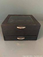 Front view of black watch and jewelry storage box with glass window on top and dual silver handles on top and drawer.