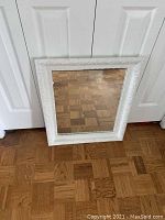 Full view of the rectangular mirror with white ornate embossed frame, showing entire mirror and frame standing against a white door on a wood parquet floor
