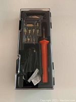 Photo of wood burning kit showing orange-handled electric tool, power cord, and eight metal tips in plastic storage case with transparent lid.