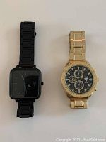 Both wristwatches shown together on a light surface, one black with square face and one gold-tone with round face and subdials.