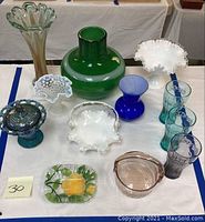 Overview of assorted glassware on white tablecloth including vases, comport, bowls, candy dish, and Coca-Cola glasses.