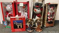 Full lot view showing two boxed plush snowman animated figures, ceramic winged angels in front, illuminated wire reindeer decoration in box, and boxed Santa Claus figure.