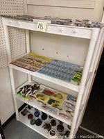 Wide view of white shelving unit stocked with multiple fishing accessories including lures in packaging, various fishing tackle items, and arranged fishing reels on the bottom shelf.