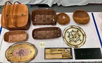 Full view of lot laid out on table showing picnic basket, woven baskets, wooden bowls, Florentine trays, and carving set in presentation boxes.