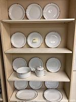 Shelves displaying white Richard Ginori dinner plates, a tea pot with lid, a serving bowl, and platters, all with red and gold geometric trim.