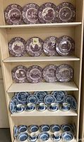 Shelved purple and blue transferware dinnerware items including plates, cups, saucers, bowls, and bread plates.