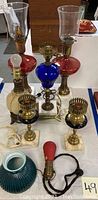 Full view of the vintage lamp lot showing two red glass hurricane lamps, blue glass lamp, brass candlestick lamps, white textured lamp base, green ribbed lamp base, and red handheld lamp bulb fixture.