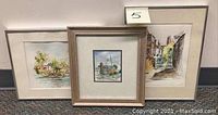 Three watercolor paintings displayed side by side with mats and frames, showing landscapes and town scenes.
