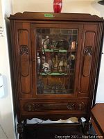 Front view of the wooden cabinet showing glass door, carved details, bottom drawer, and cabinet legs.