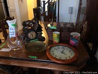 Photo shows glass decanter, mantel clock with horse figure, round wall clock, metal bowl, and two decorative candles on wooden table.