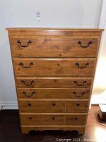 Front view of a tall wooden dresser with 5 drawers and black metal handles, showing wood grain and knots.