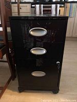 Front view of black metal filing cabinet with three drawers, showing oval silver handles and some surface scratches.