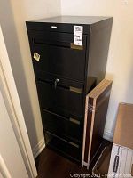Full view of the black metal filing cabinet showing four drawers with labels and key lock on one drawer.
