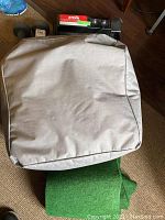 Gray cushioned carrying case for indoor electric golf putting green mat on brown floor with partial green mat visible underneath