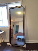 Full-frontal view of the large framed mirror leaning against a wall by a window.