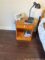 Wooden bedside table with drawer, white knob, open shelf, located beside a bed, with small electronic devices and a lamp on top