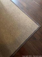 Close-up image of the corner of a large rectangular rug showing woven texture and dark brown border on hardwood floor.