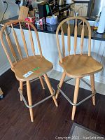 Two wooden bar stools with spindle backs and metal foot bars, positioned in kitchen area showing moderate wear and scratches on wood surfaces.