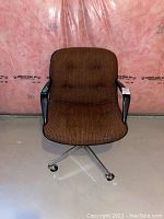 Vintage office chair with brown patterned fabric upholstery, black armrests, metal swivel base, and casters against a basement wall.