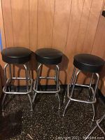 Three swivel barstools with black vinyl seats and chrome frames against wood panel wall.