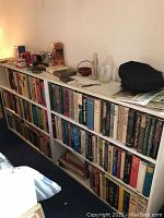 White bookshelf filled with various hardcover and softcover books with some miscellaneous items on top.