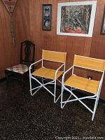 Pair of folding director's chairs with yellow fabric seats and backs, white metal frames, placed against wood-paneled wall with artwork in background.
