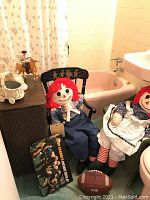 Wooden child rocker painted black with decorative design, Raggedy Ann and Andy dolls seated on it, leather football, horse rider figurine on the wicker basket planter beside a green wicker rocker chair.