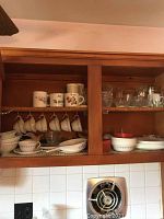 Cabinet shelf showing vintage CorningWare cups, mugs, saucers, bowls, and additional glassware.