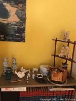 Wide view showing multiple porcelain figurines, jewelry box, deel tray, and ashtray on shelves against yellow wall