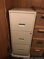 Front view of beige metal file cabinet showing all three drawers, middle drawer with key lock. Cabinet positioned against wooden wall next to stacked cardboard boxes.
