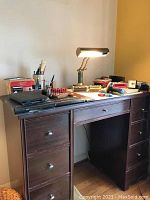 Dark wooden desk with bankers lamp, clocks, calculator, label maker, stapler, and other office supplies.