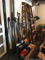 Full view of the lot including shovels, rakes, step ladders, and a Black & Decker electric shrub trimmer on the floor.