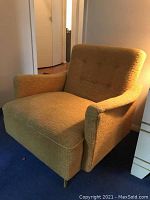 Front angled view of a gold/yellow upholstered mid century modern armchair on blue carpet, showing armrests and button tufting on backrest.