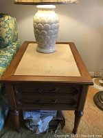 Overall view of one end table with a ceramic lamp on top, showing the wooden frame and drawer fronts below the stone top insert.