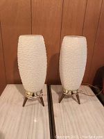 Two mid century modern white textured glass lamps with brass rings and tripod wooden bases shown side by side