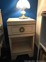 Mid century modern nightstand shown with lamp on top, white paint with visible wear and circular drawer pull.