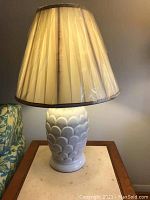 Photo of one table lamp showing ceramic base with scalloped scale pattern and beige pleated lamp shade wrapped in plastic.