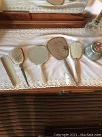 Photo showing the vintage vanity set laid out on a lace cloth including hand mirror, brushes, combs, and powder jar.