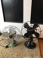 Two vintage table fans side-by-side on a carpeted floor in front of a black and white door, one black Westinghouse Whirlwind fan and one green fan with clear plastic blades.